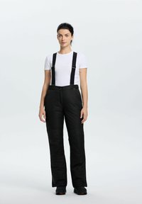 Kobieta stoi przodem, ubrana w białą koszulę z krótkim rękawem, czarne ocieplane spodnie śnieżne z szelkami oraz czarne zimowe buty na jednolitym tle.