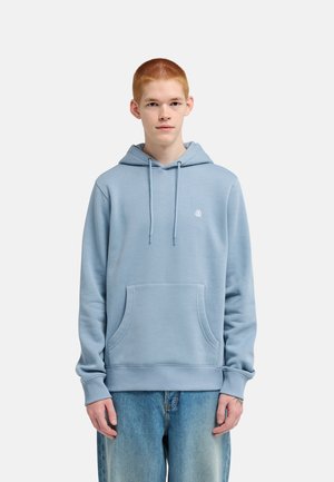 Persona joven con cabello corto y rojo, vistiendo una sudadera con capucha azul claro y vaqueros azules, de pie frente a un fondo blanco liso.