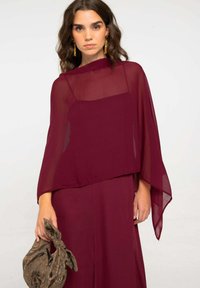 Top in chiffon marrone con un design a strati, fluido e con maniche ampie; abbinato a una pochette marrone con motivo.