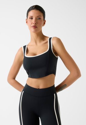 Femme portant un crop top noir avec une bordure blanche et des leggings noirs avec des rayures blanches, debout, les bras derrière le dos.
