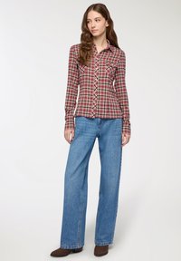 Donna in piedi, che indossa una camicia a quadri rossa e bianca, jeans larghi blu e stivali marroni a punta, su uno sfondo bianco.