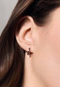 Złota kolczyk w kształcie obręczy wyeksponowany na uchu, z wypolerowaną powierzchnią i gładkim, zaokrąglonym kształtem. Włosy są stylizowane z dala od ucha.