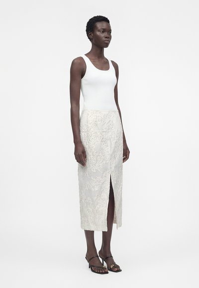 Γυναίκα που στέκεται φορώντας ένα λευκό αμάνικο ριμπ top και μια λευκή υφασμάτινη φούστα μέχρι το γόνατο με μπροστινή σχισμή, συνδυασμένη με μαύρα σανδάλια με τακούνι.