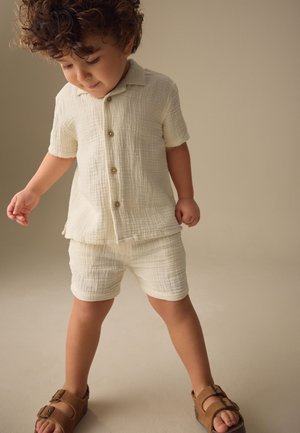 Kleinkind mit lockigem Haar, das ein off-white strukturiertes kurzärmeliges Hemd mit Knöpfen und passende Shorts trägt, steht mit gespreizten Füßen in braunen Sandalen.