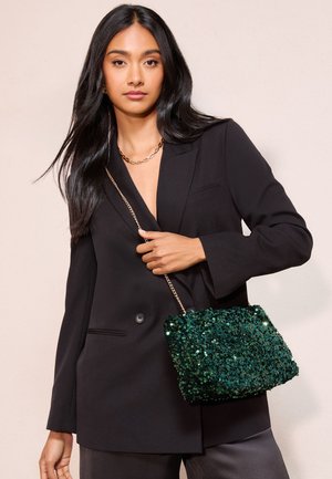 Donna con capelli lunghi e neri che indossa un blazer nero e una collana a catena, mentre tiene una borsa a tracolla verde con paillettes e una catenella dorata.