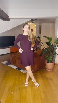 Ung kvinne med langt hår i lilla kjole og hvite sandaler står med hendene på hoftene i en stue med planter og en TV.