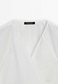 Top enveloppant blanc en tissu léger, avec un décolleté en V profond, des manches larges et une fermeture éclair au dos. Étiquette visible au niveau du col.