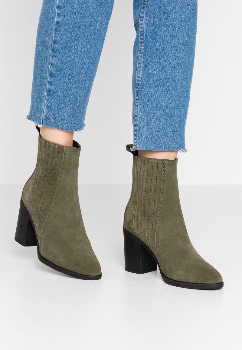 Bottes en daim vert olive longueur cheville avec talons carrés portées avec un jean bleu à ourlet effiloché sur une surface blanche.