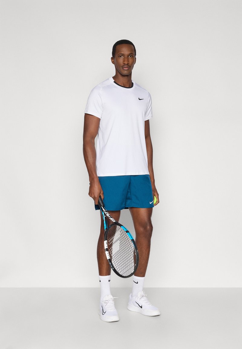 Vit Nike T-shirt med svart detalj, blå shorts, vita tennisskor, håller i ett tennisracket och en boll, mot en enfärgad bakgrund.