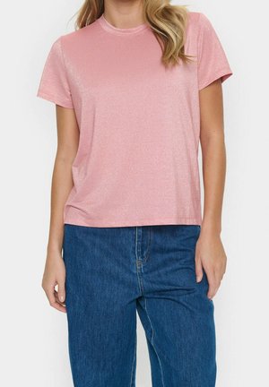 Femme portant un t-shirt à manches courtes, rose clair et brillant, et un jean bleu en denim ample, debout devant un fond blanc.