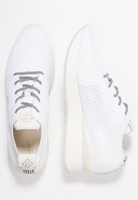 Baskets blanches en maille avec dessus texturé, lacets à motifs noirs et blancs, et semelle blanche. Marque visible sur le talon.