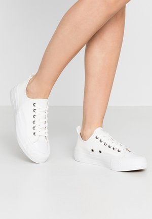 Ben iført hvide lave snørestøvler med tekstureret gummisål på en lys grå gulv og baggrund.