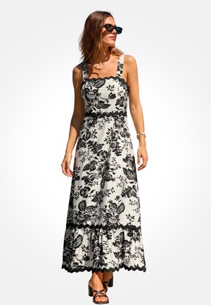 Maxi-jurk in wit met zwart bloemenpatroon, vierkante halslijn, brede bandjes en gerimpelde zoom. Draagt zwarte sandalen en een zonnebril.