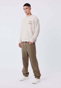 Giovane uomo in piedi che indossa un maglione beige con logo di aereo, pantaloni marroni a fantasia e sneakers bianche su uno sfondo neutro.