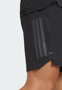 Pantalones cortos deportivos negros hechos de un material ligero, con tres rayas reflectantes verticales en el lateral y un bolsillo con cremallera lateral.
