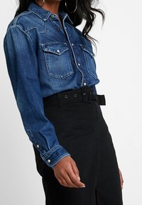Chemise en denim à boutons, avec deux poches poitrine, boutons-pression et manches longues, assortie d'une jupe noire taille haute avec une ceinture.