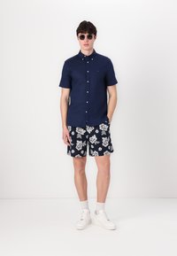 Marineblå, kortermet knappeopp skjorte, marineblå shorts med blomstermønster, hvite sneakers og solbriller; avslappet sommerantrekk med kontrastfarger.
