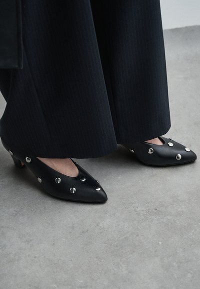 Črne usnjene pete s koničastim prstom, ki imajo srebrne žeblje, nizko blok peto in gladko teksturo, v kombinaciji s širokimi črnimi hlačami s črtami.