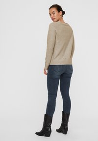 Lys beige sweater med ribbet kant og manchetter, parret med mørkeblå tætsiddende jeans og sorte stilethæle, vist fra ryggen.