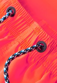 Neon oranje zwembroek met een zwarte grommet waarop "SUPERDRY" is geperst, en een textuurkoord met blauwe en witte accenten.