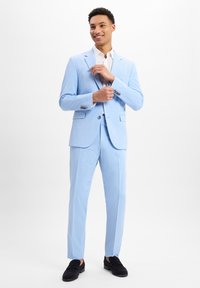 Lichtblauw pak van gestructureerde stof, enkelknops jasje, bijpassende broek en zwarte instappers. Witte overhemd eronder.