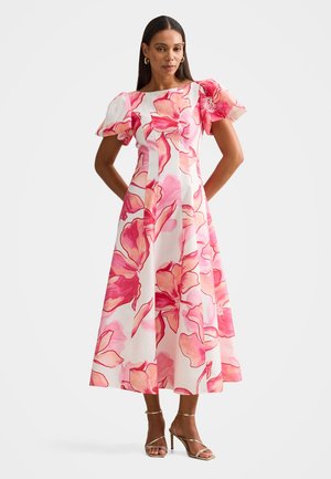Donna con un vestito midi bianco con grandi stampe floreali rosa e maniche a sbuffo, abbinato a sandali dorati con tacco alto.