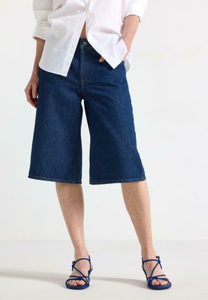 Kvinde iført mørkeblå knee-length cowboyshorts, en hvid skjorte med knapper og blå sandaler med hæl og stropper.