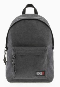 Mochila de tela gris con parte superior redondeada, bolsillo frontal con cremallera, correas ajustables, acentos negros y una etiqueta de logo en la parte delantera.