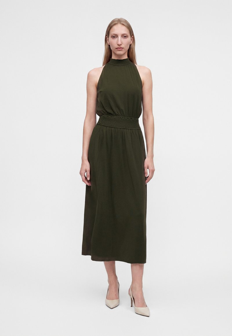 Femme debout portant une robe midi sans manches vert foncé à col montant et des chaussures à talons beiges sur un fond uni.