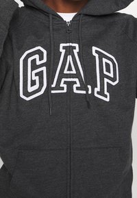 Sudadera de color gris carbón con un gran logo blanco de "GAP", cierre de cremallera, capucha con cordón y bolsillos frontales, fabricada con una tela suave y texturizada.