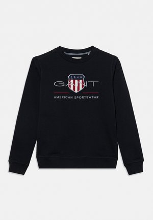 Sweat-shirt noir à manches longues avec col rond, arborant un logo bouclier rouge et blanc et le texte "GANT 1949 American Sportswear" sur le devant.