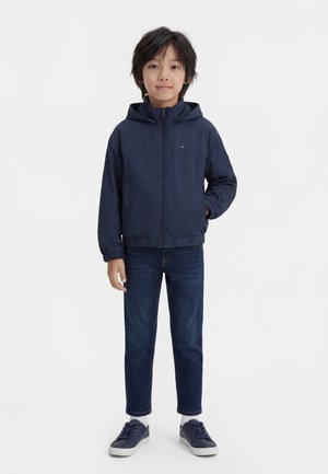 Junge steht mit dem Gesicht nach vorne und den Händen teilweise in den Taschen, trägt eine marineblaue Kapuzenjacke, dunkle Jeans, weiße Socken und marineblaue Turnschuhe.