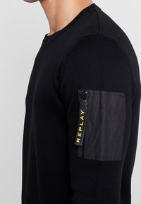Homme portant un pull en tricot noir avec une poche zippée sur le bras comportant une étiquette noire avec le mot "REPLAY" en lettres jaunes.
