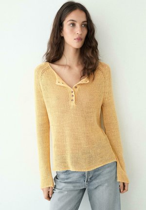 Vrouw met lang bruin haar, gekleed in een geel gebreid shirt met lange mouwen en knopen, en lichtblauwe spijkerbroek, tegen een witte achtergrond.