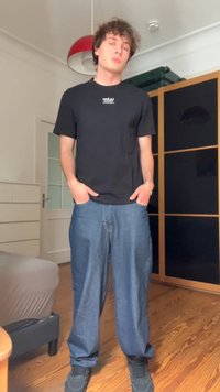 Černé tričko s kulatým výstřihem a malým bílým logem, v kombinaci s volnými modrými džíny. Oblečení dotvářejí tmavé tenisky. Pozadí tvoří dřevěná podlaha.