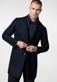 Granatowy wełniany płaszcz o dopasowanym kroju, z klapami i dwoma guzikami. Noszony na niebieskim golfie, z teksturowanym wewnętrznym kołnierzem.