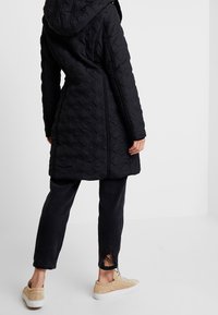 Černý prošívaný kabát s kapucí, který má geometrický vzor. Nosený s tmavými džíny s třepenými lemy a béžovými teniskami.