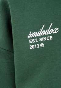 Groene stof met witte tekst "smilodox EST. SINCE 2013 ©" erop gedrukt, waarschijnlijk onderdeel van een kledingstuk.