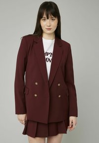 Blazer bordeaux à double boutonnage avec des boutons dorés, doté d'un col cranté et de poches latérales, associé à une mini-jupe plissée.
