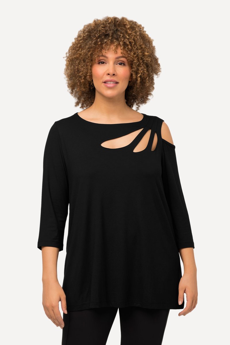Femme aux cheveux bouclés portant un haut noir à manches longues avec des découpes près de l'épaule droite et du décolleté, debout devant un fond uni.