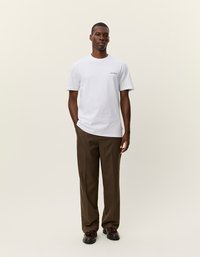 Λευκό μπλουζάκι με στρογγυλή λαιμόκοψη και μικρό μαύρο λογότυπο στο αριστερό στήθος, συνδυασμένο με καφέ καρό παντελόνι και μαύρα παπούτσια. Απλή, casual εμφάνιση.