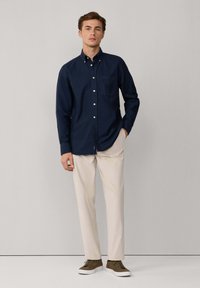 Camisa de botón azul marino con un bolsillo en el pecho, combinada con pantalones beige y zapatillas oliva, contra un fondo neutro.
