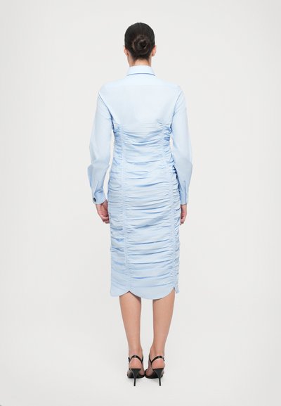 Rochie bleu deschis, cu detalii plisate, spate elaborat, maneci lungi, guler și tiv până la genunchi. Asortată cu sandale negre cu barete.