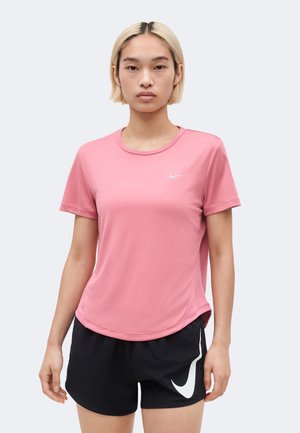 Vrouw met kort blond haar, gekleed in een roze Nike T-shirt met korte mouwen en zwarte Nike shorts, staand tegen een effen lichte achtergrond.