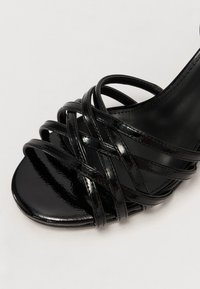 Sandal cu toc din piele patent neagră, cu mai multe curele încrucișate și un design cu vârful deschis. Suprafață netedă și lucioasă, cu o talpă conturată.
