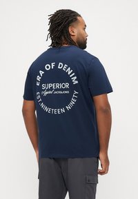 Tamsiai mėlyna medvilninė marškinėliai trumpomis rankovėmis. Ant nugaros baltomis raidėmis užrašyta „ERA OF DENIM“, „SUPERIOR“ ir „EST. NINETEEN NINETY“.