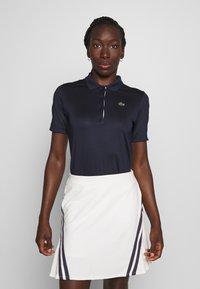 Polo shirt navy con maniche corte, dettagli bianchi e un piccolo logo verde; abbinato a una skort bianca con strisce laterali scure.