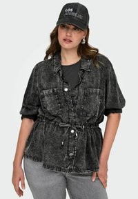 Črna kislo oprana jeans srajca z gumbnim zapiranjem, dvema žepoma na prsih, elastičnim pasom in zavihanimi rokavi, v kombinaciji s črno kapo.