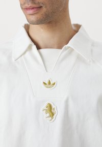 Bílá košilová košile s vyšitým zlatým logem Adidas a zlatým zvířecím emblémem, vyrobená z texturované bavlny.