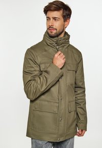 Olivenfarvet jakke med høj krave, lynlås- og knaplukninger, to forlommer og et glat, struktureret stof. Casual pasform.
